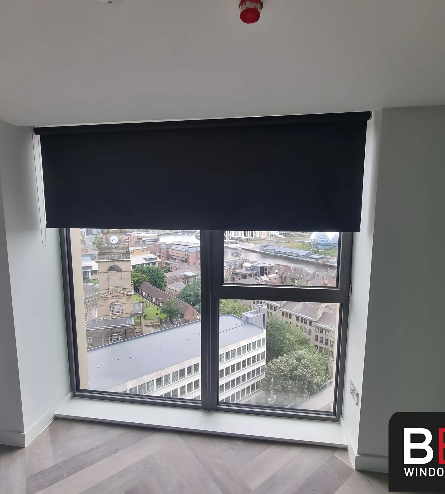 A dark blackout roller blind half lowered over a bedroom window with the city below
