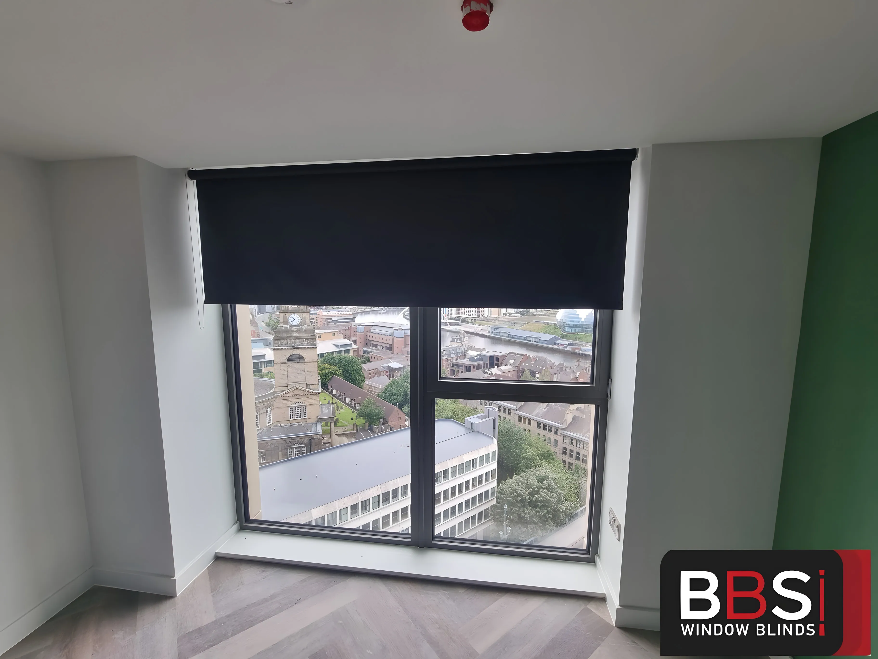 A dark blackout roller blind half lowered over a bedroom window with the city below