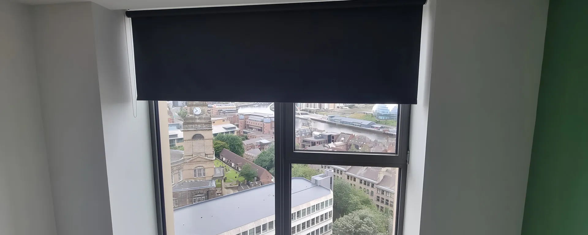 A dark blackout roller blind half lowered over a bedroom window with the city below