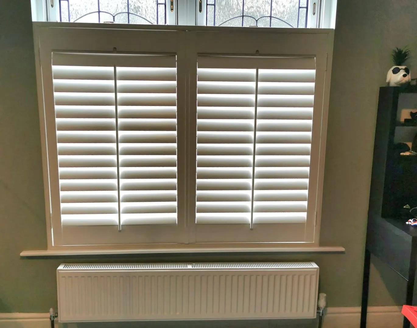 White café style shutters on the lower half of a period window, with the original stained glass lights left clear above