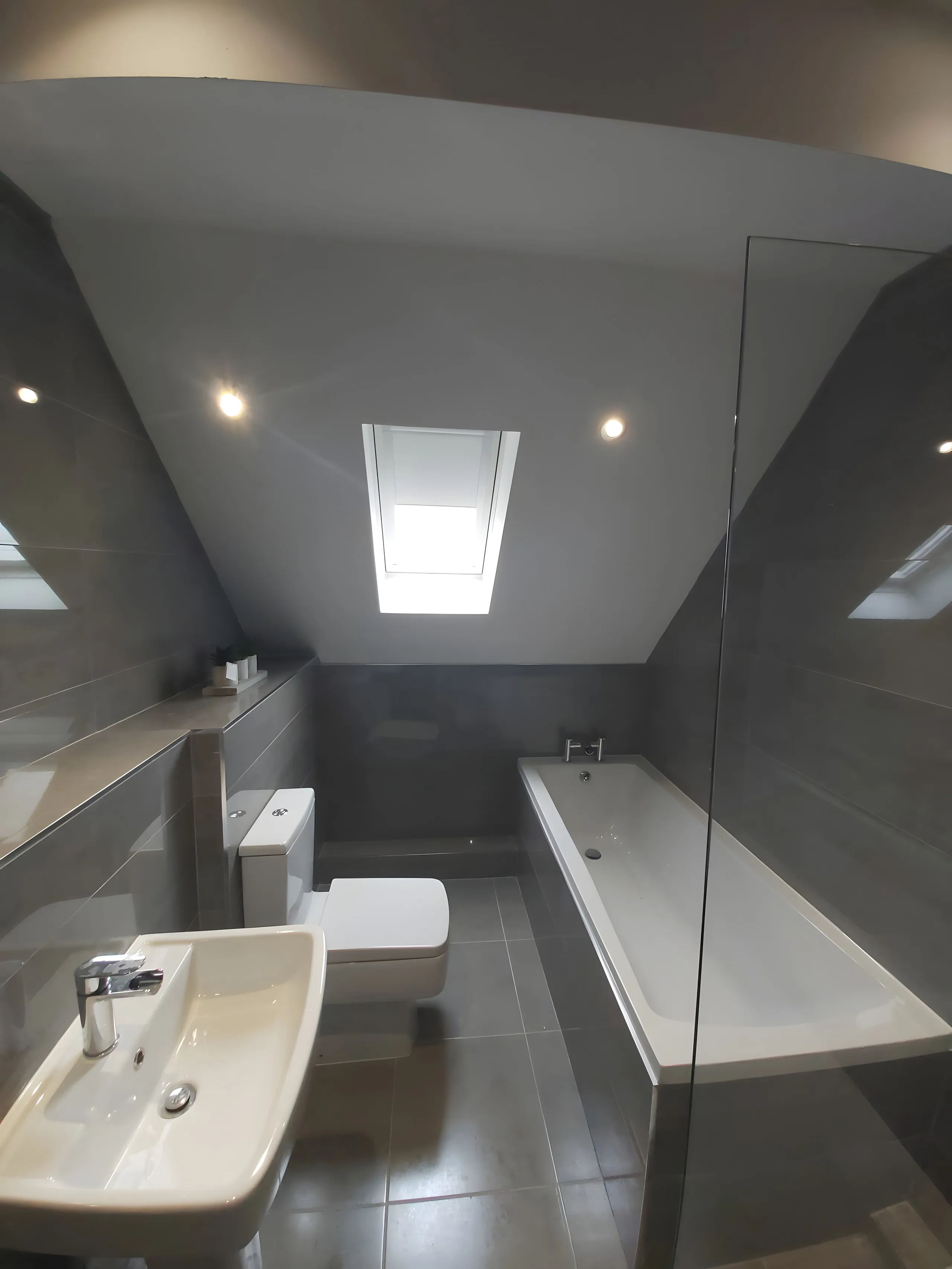 Roof window blind fitted above the bath in a grey-tiled loft bathroom