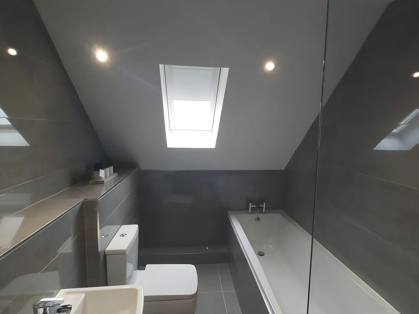 Roof window blind fitted above the bath in a grey-tiled loft bathroom