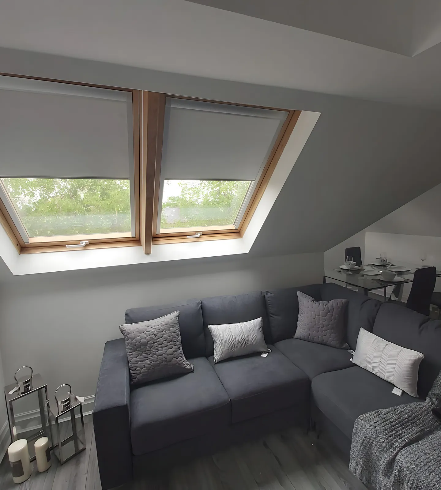 Two roof window blinds fitted in a loft living room above a grey corner sofa