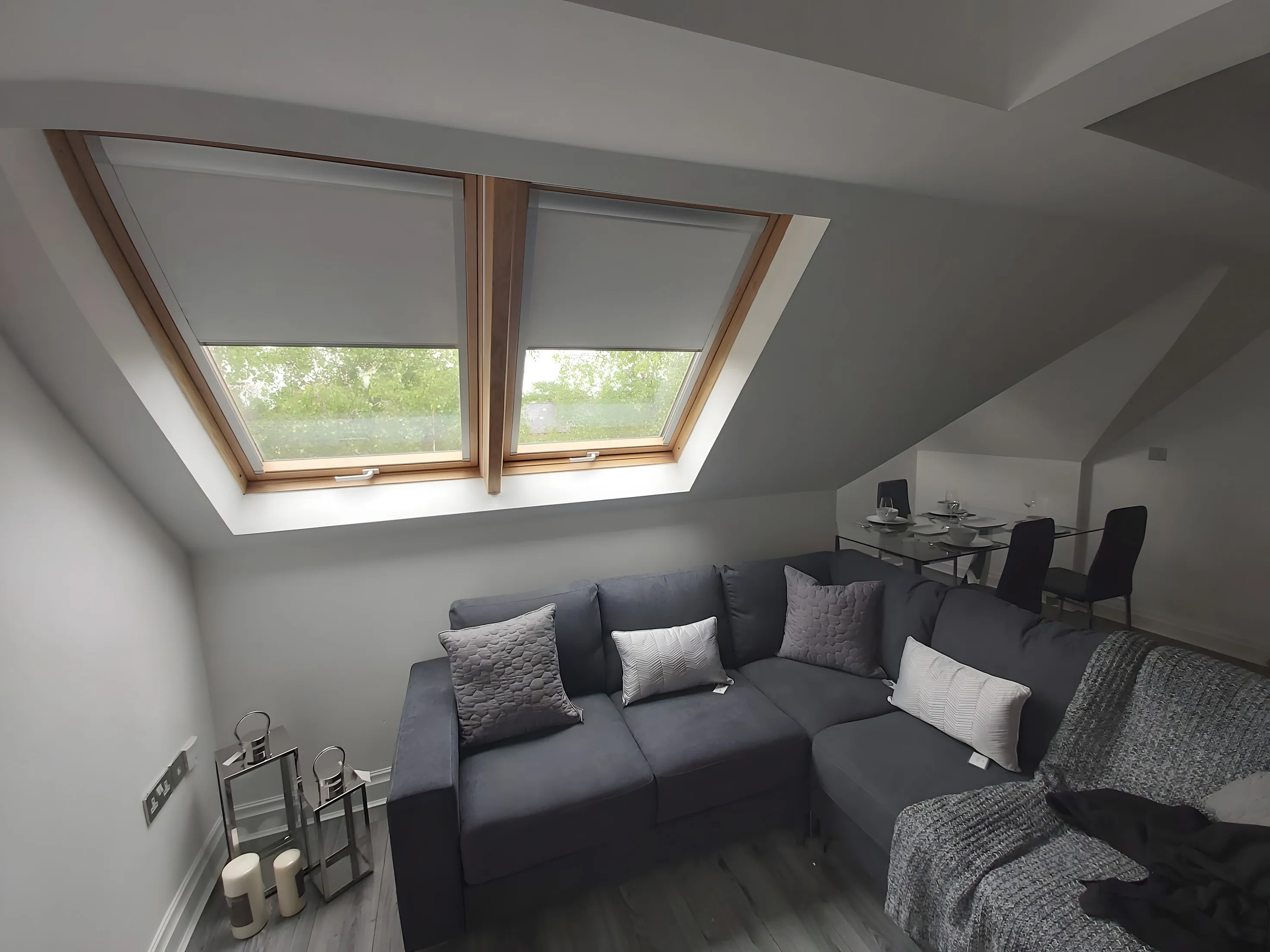 Two roof window blinds fitted in a loft living room above a grey corner sofa