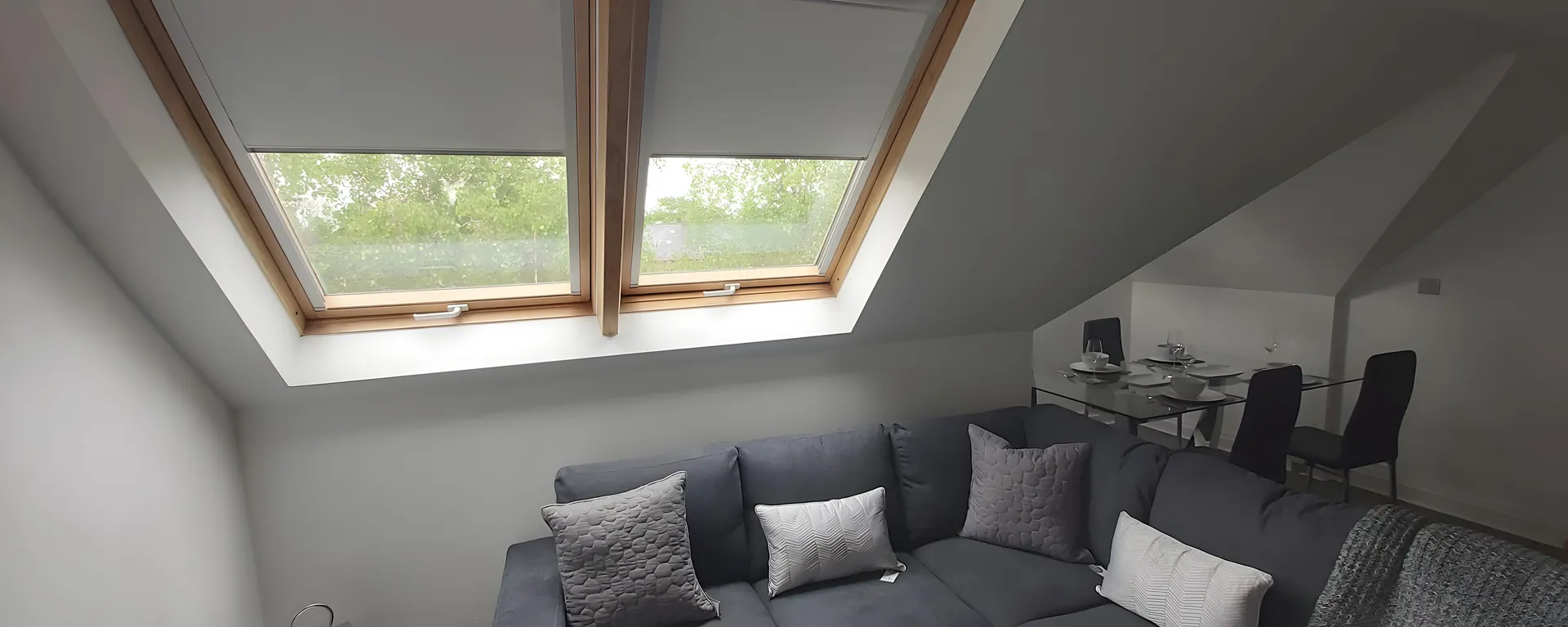 Two roof window blinds fitted in a loft living room above a grey corner sofa