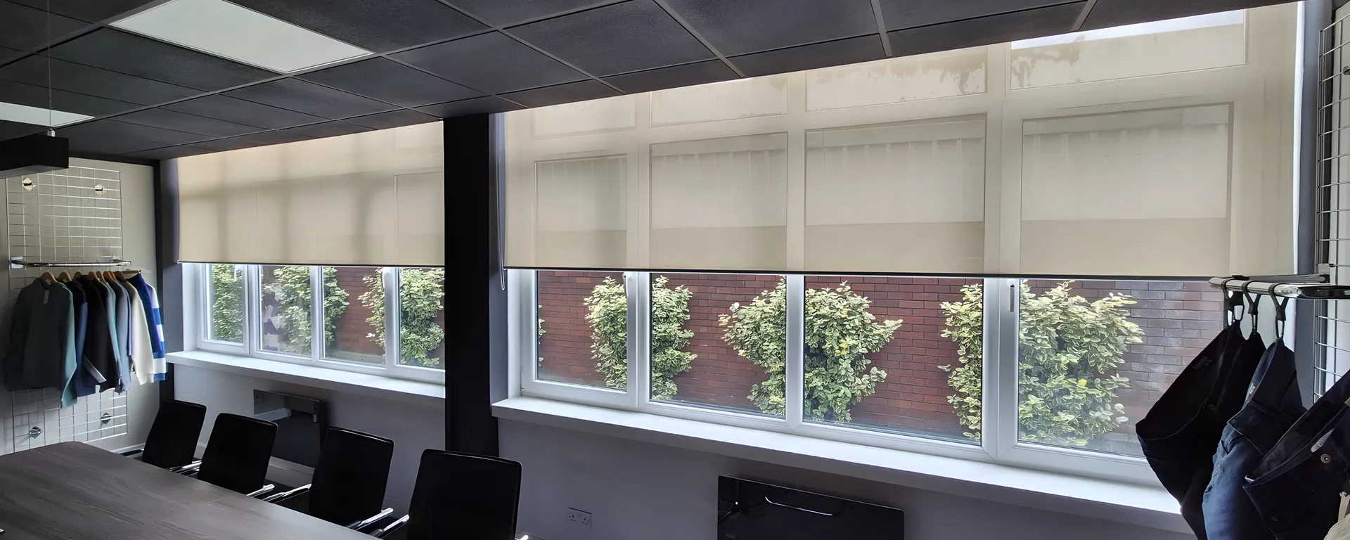 Boardroom with five screen fabric roller blinds set to a matching half drop along a full width window, greenery and brickwork visible through the fabric