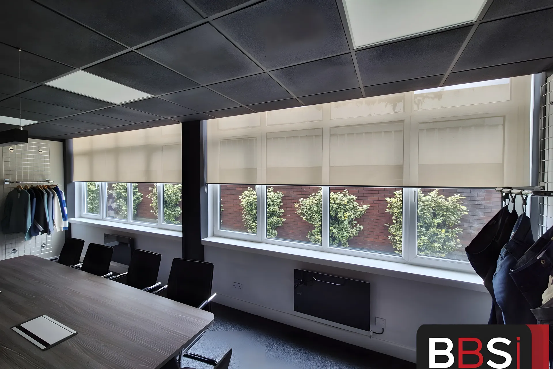 Boardroom with five screen fabric roller blinds set to a matching half drop along a full width window, greenery and brickwork visible through the fabric