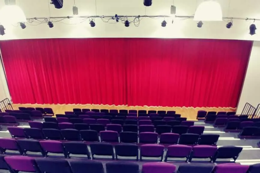 Full-width red stage curtain across a school hall, rows of purple tiered seating in front