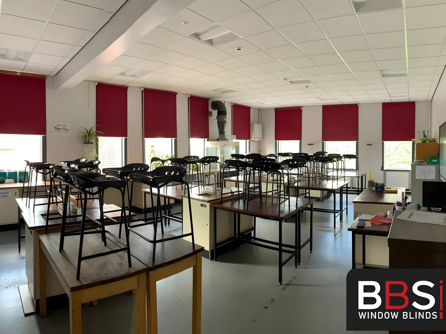 Burgundy roller blinds across tall Stockport School classroom windows, with lab stools stacked on the desks