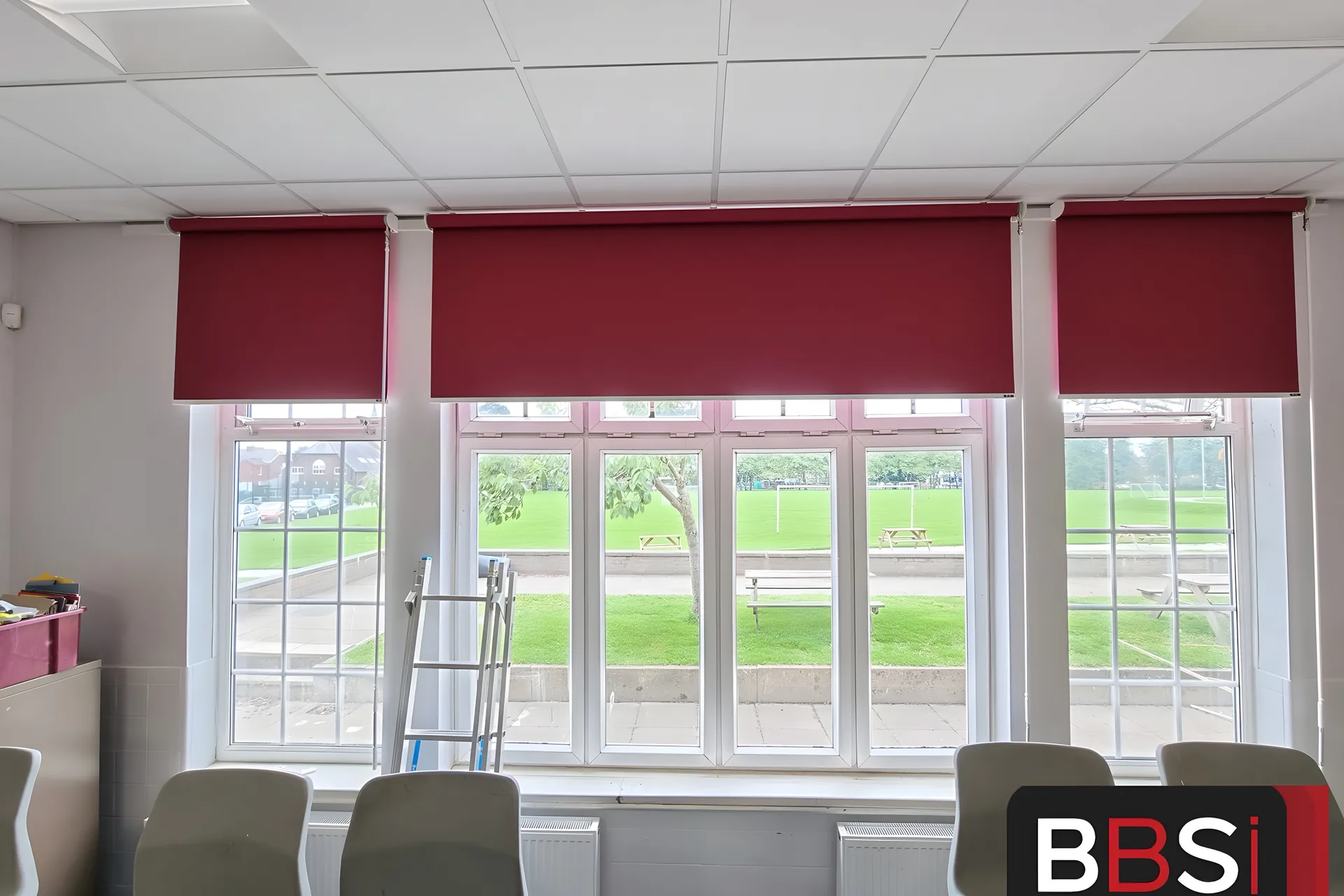 Three burgundy roller blinds fitted above a bay of tall white sash windows in a Stockport School classroom, playing field beyond