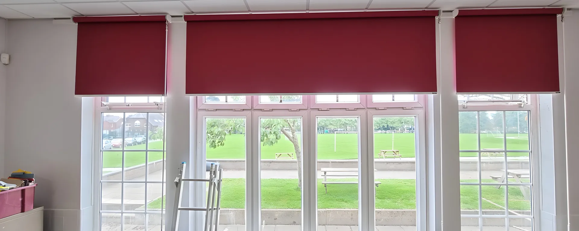 Three burgundy roller blinds fitted above a bay of tall white sash windows in a Stockport School classroom, playing field beyond