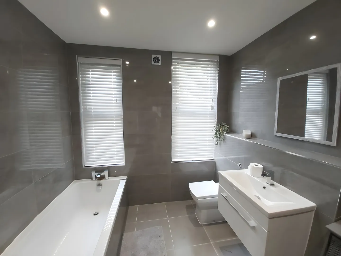 White Venetian blind at the window of a grey-tiled bathroom above the bath