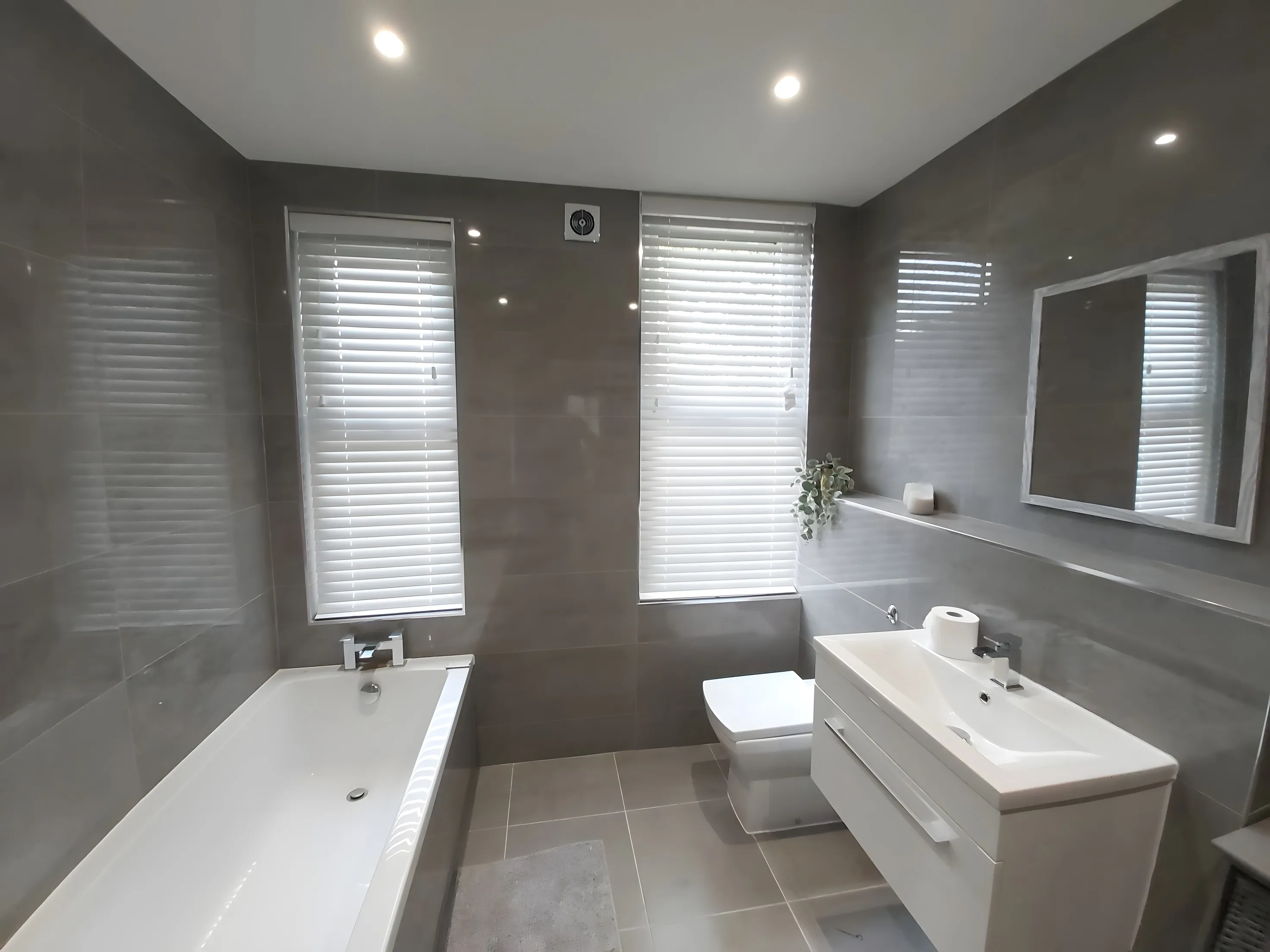 White Venetian blind at the window of a grey-tiled bathroom above the bath