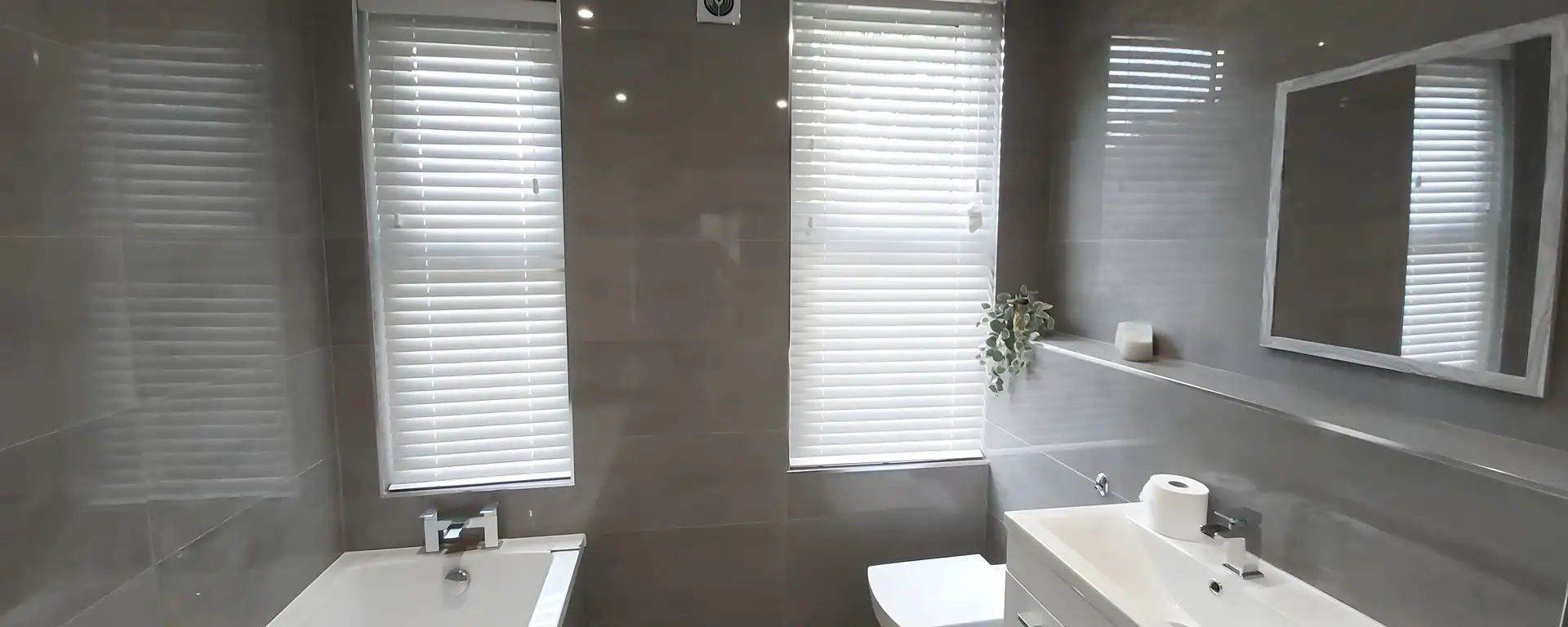 White Venetian blind fitted to the window of a grey-tiled bathroom above the bath