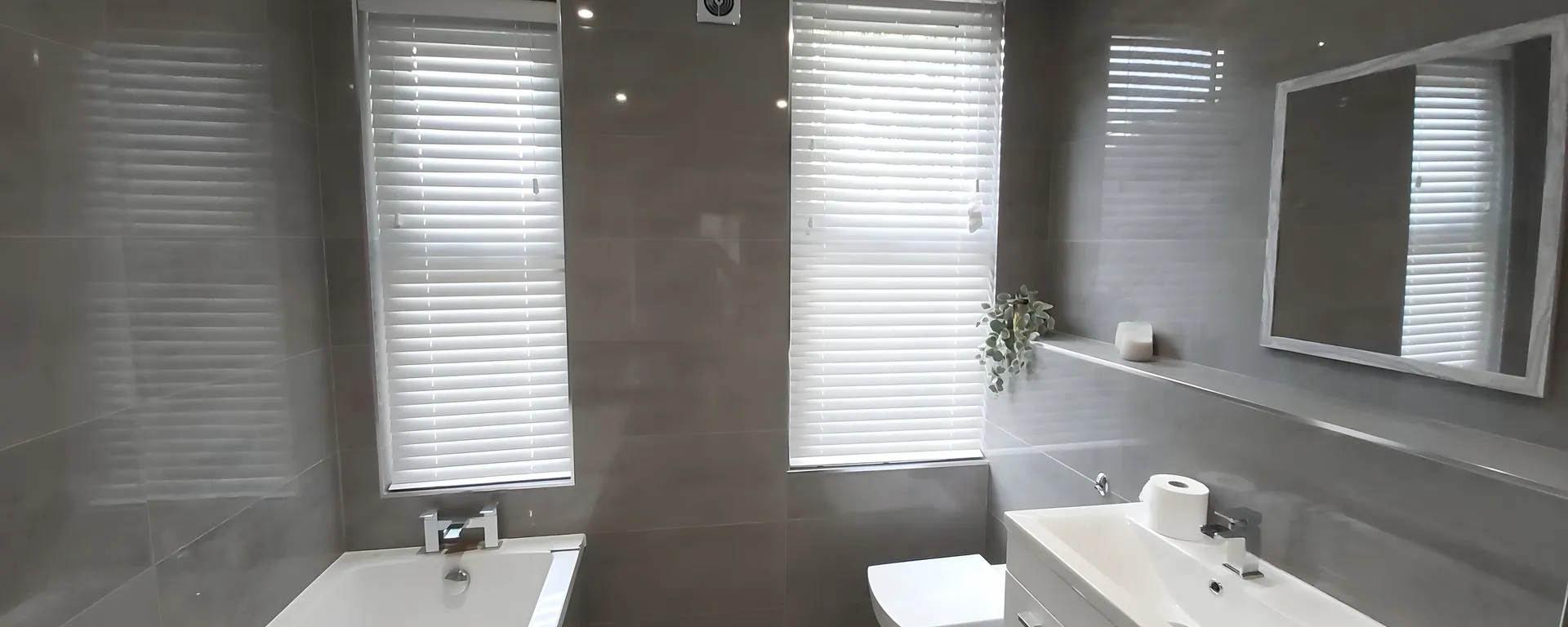 White Venetian blind fitted to the window of a grey-tiled bathroom above the bath