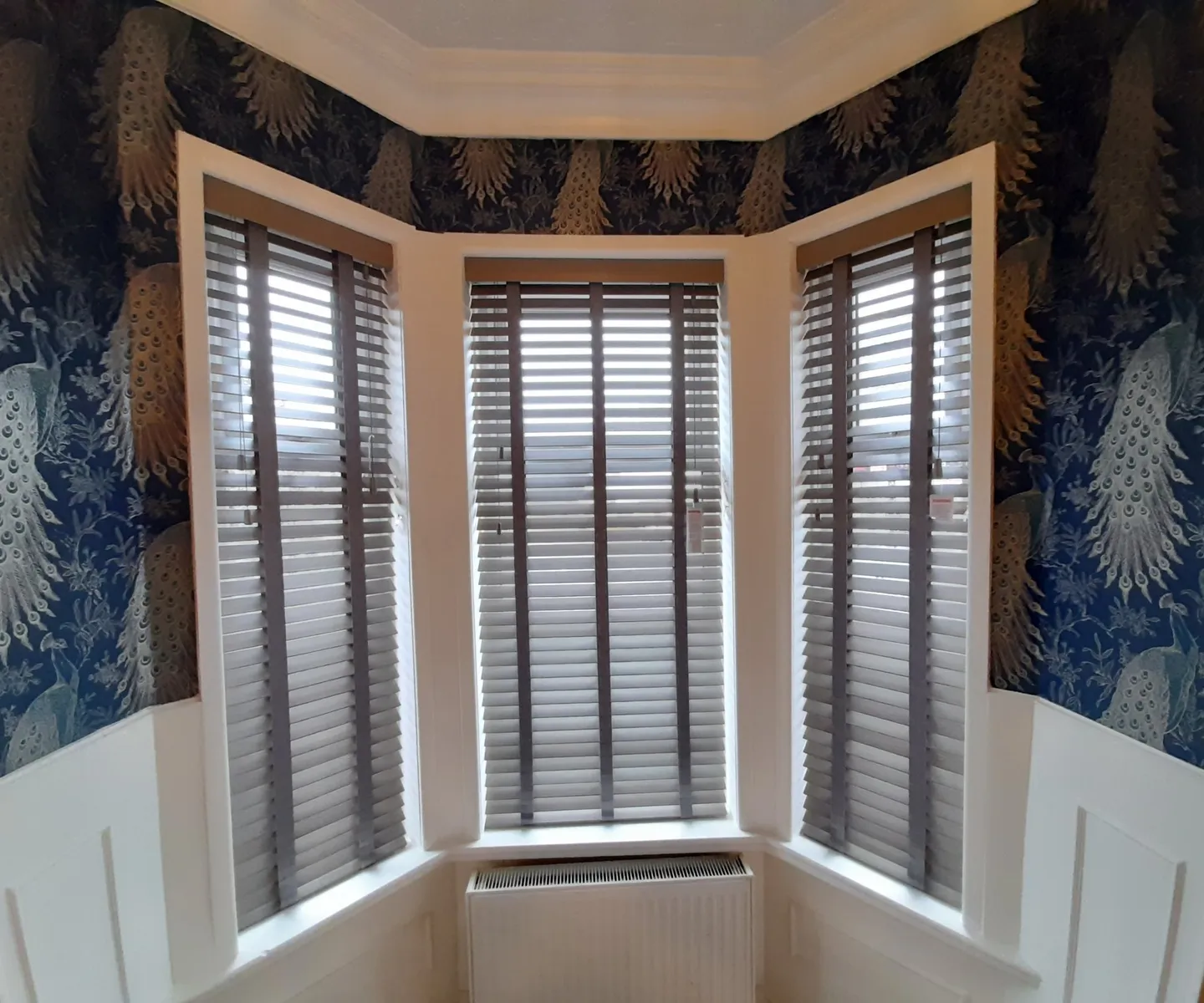 White wooden Venetian blinds in a bay window framed by dark botanical wallpaper