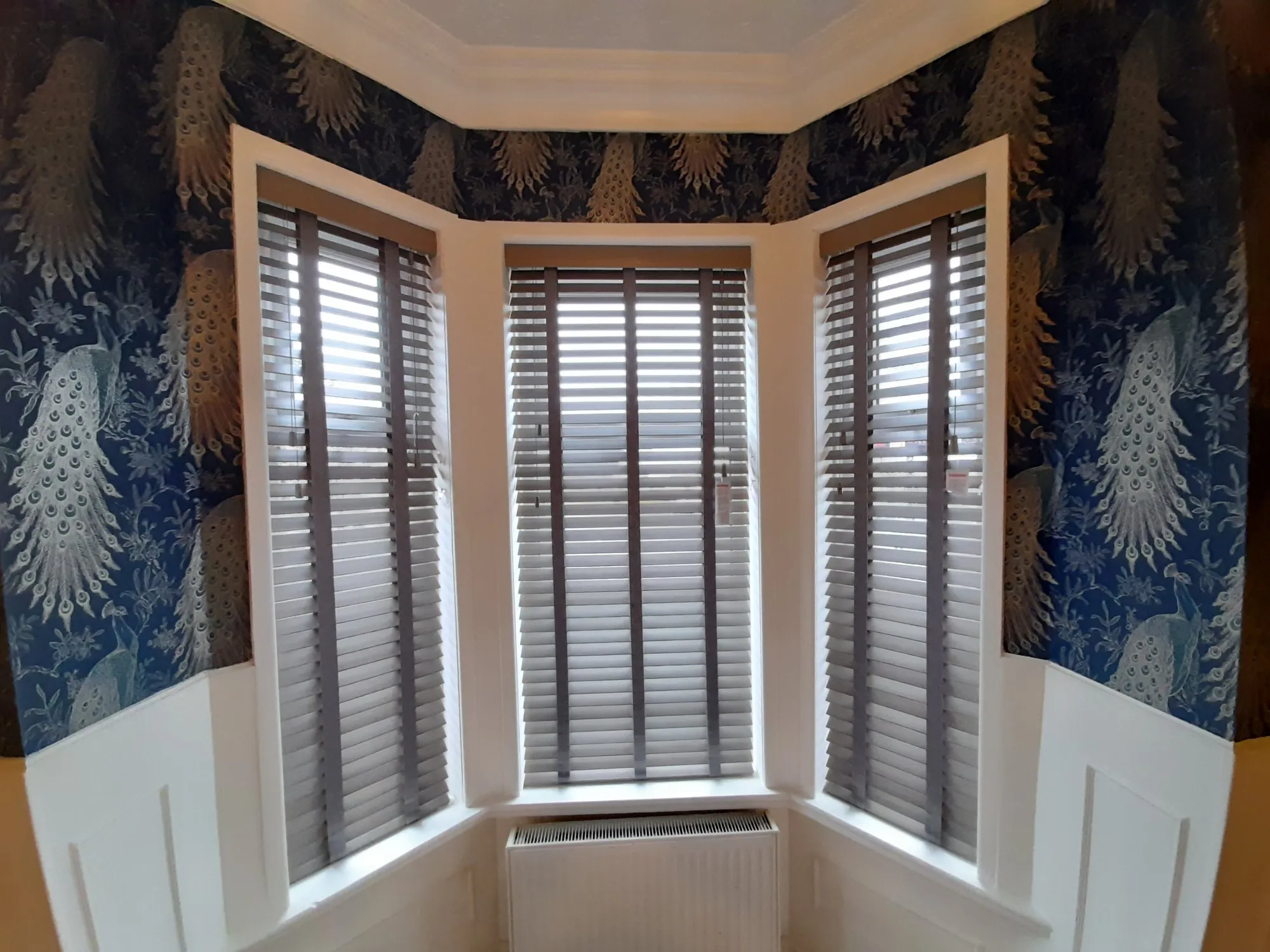 White wooden Venetian blinds in a bay window framed by dark botanical wallpaper