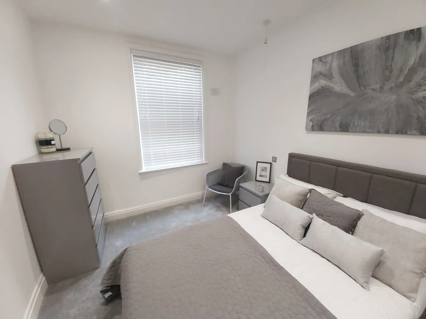 White Venetian blind at a bedroom window with grey walls and a made-up bed