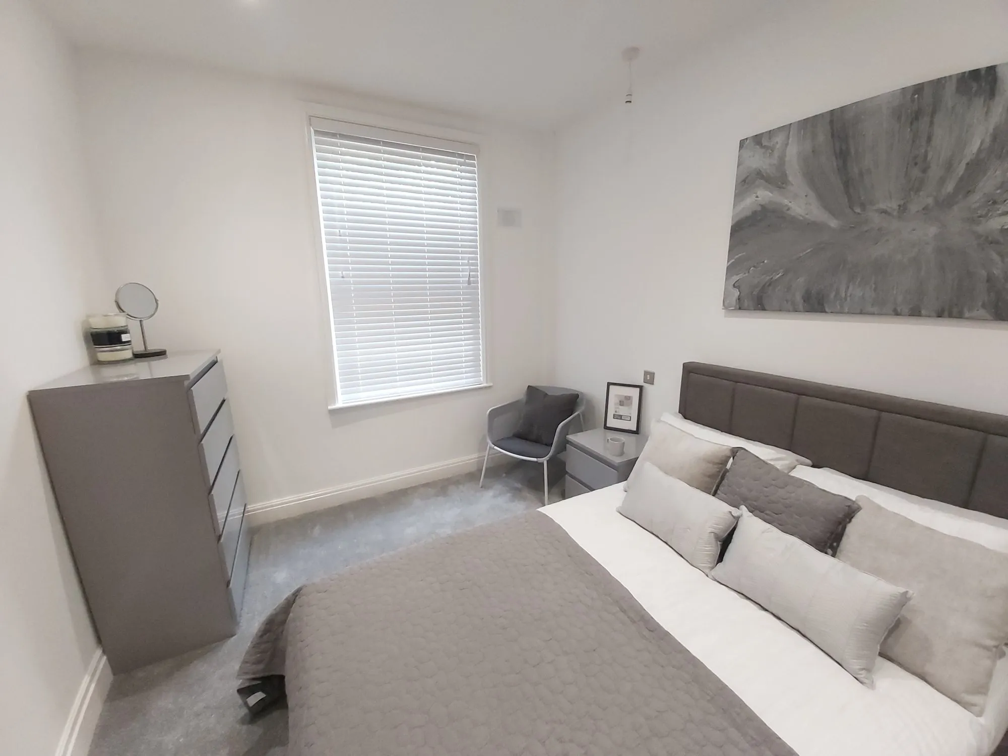 White Venetian blind at a bedroom window with grey walls and a made-up bed