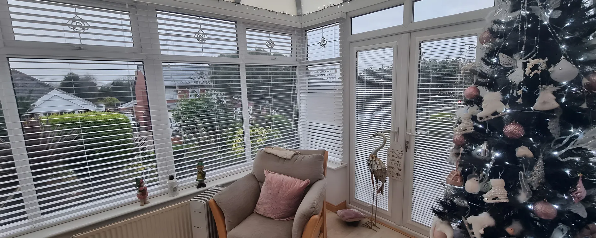White Venetian blinds fitted to every window of a conservatory with a Christmas tree and armchair