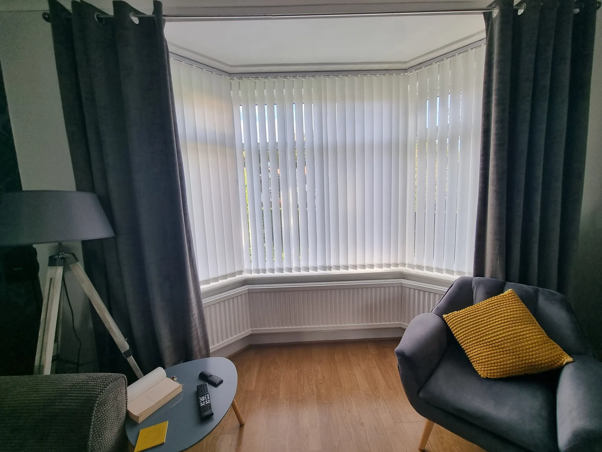 White vertical blinds across a bay window framed by charcoal eyelet curtains on a pole, with an armchair and yellow cushion