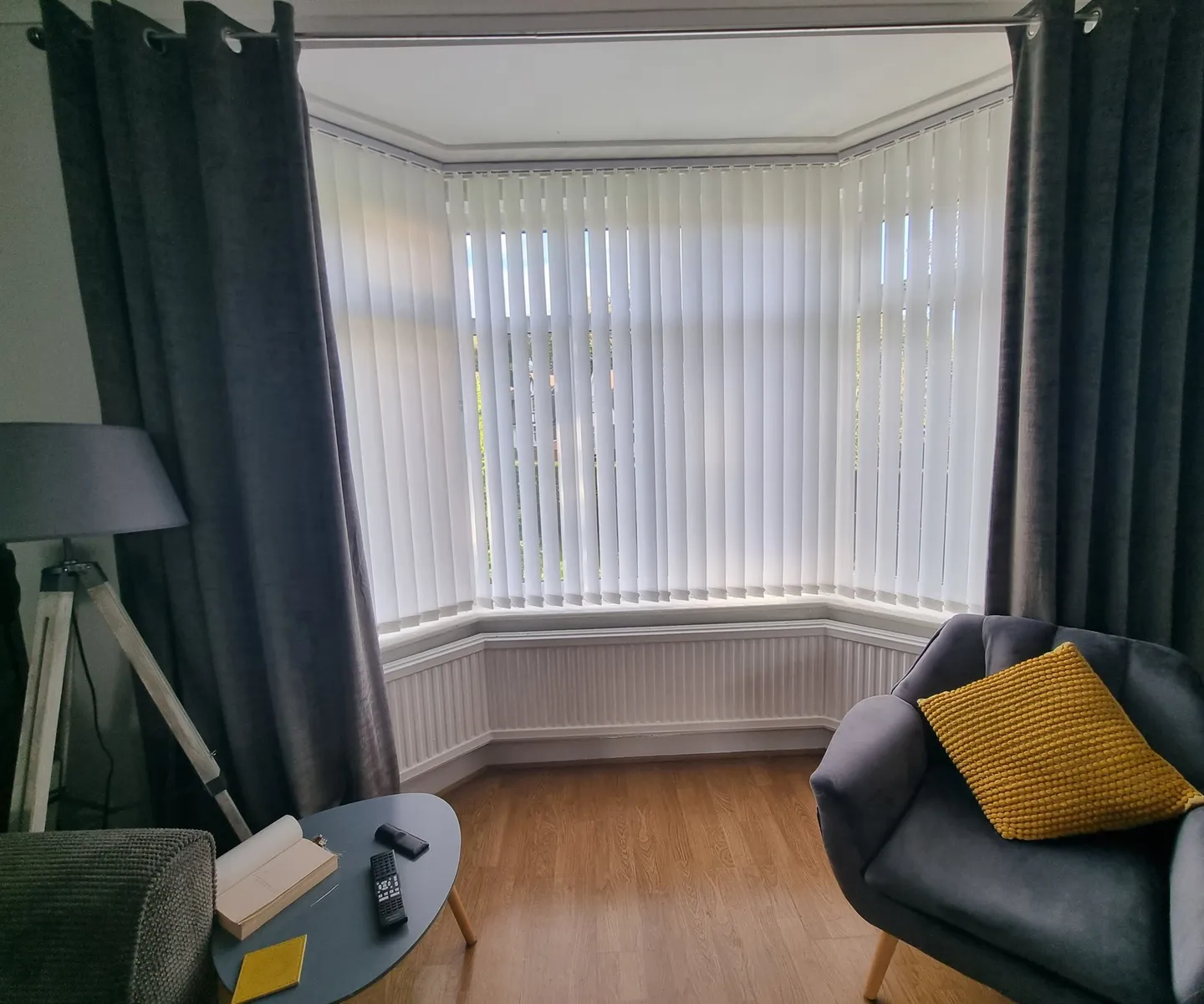 Cream vertical blinds inside a bay window with grey curtains either side and a yellow cushion on the chair