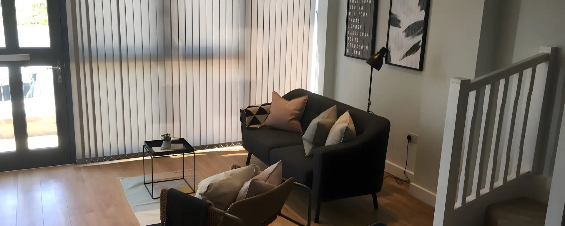 Grey vertical blinds across full-height glazing in a living room with a dark sofa and armchairs