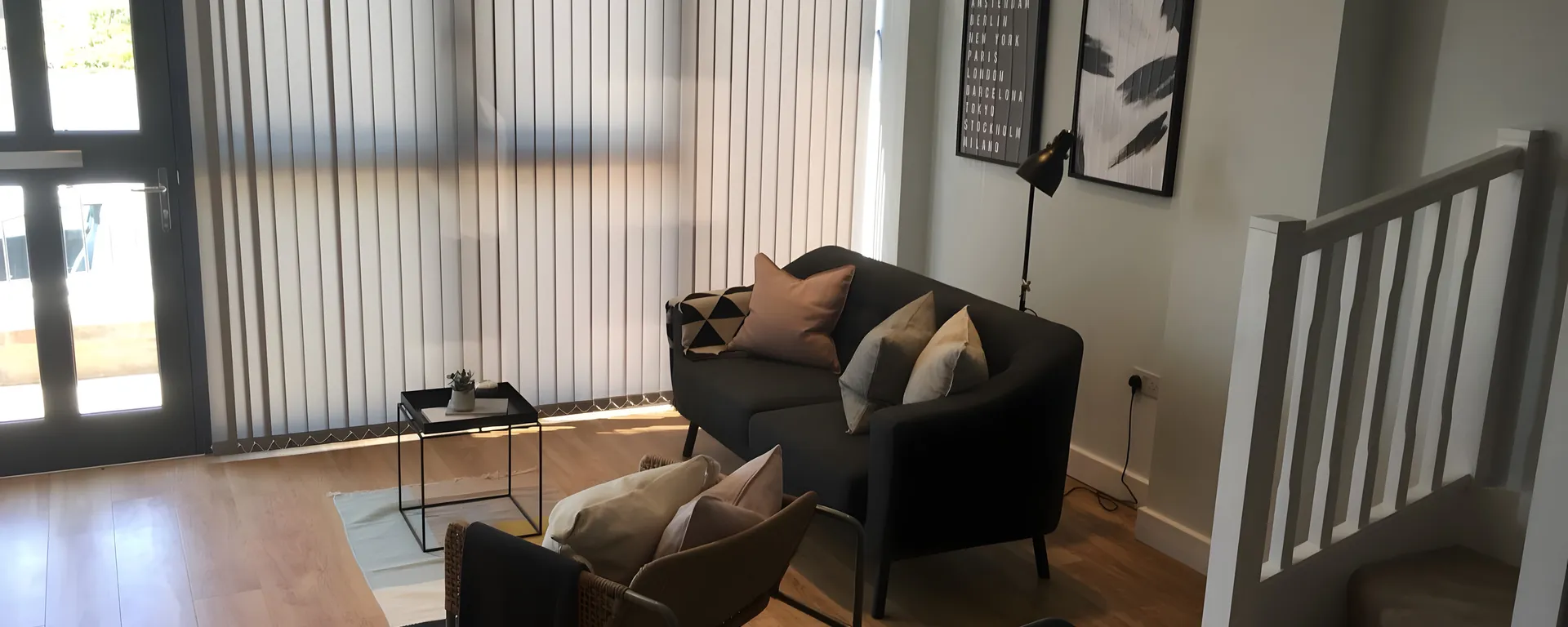 Grey vertical blinds across full-height glazing in a living room with a dark sofa and armchairs